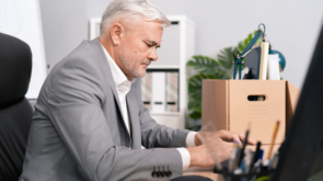 Focused elderly company executive sits in front of computer monitor taps fingers on keyboard looks at buttons carefully elegant man with gray hair spends last day at work before retirement