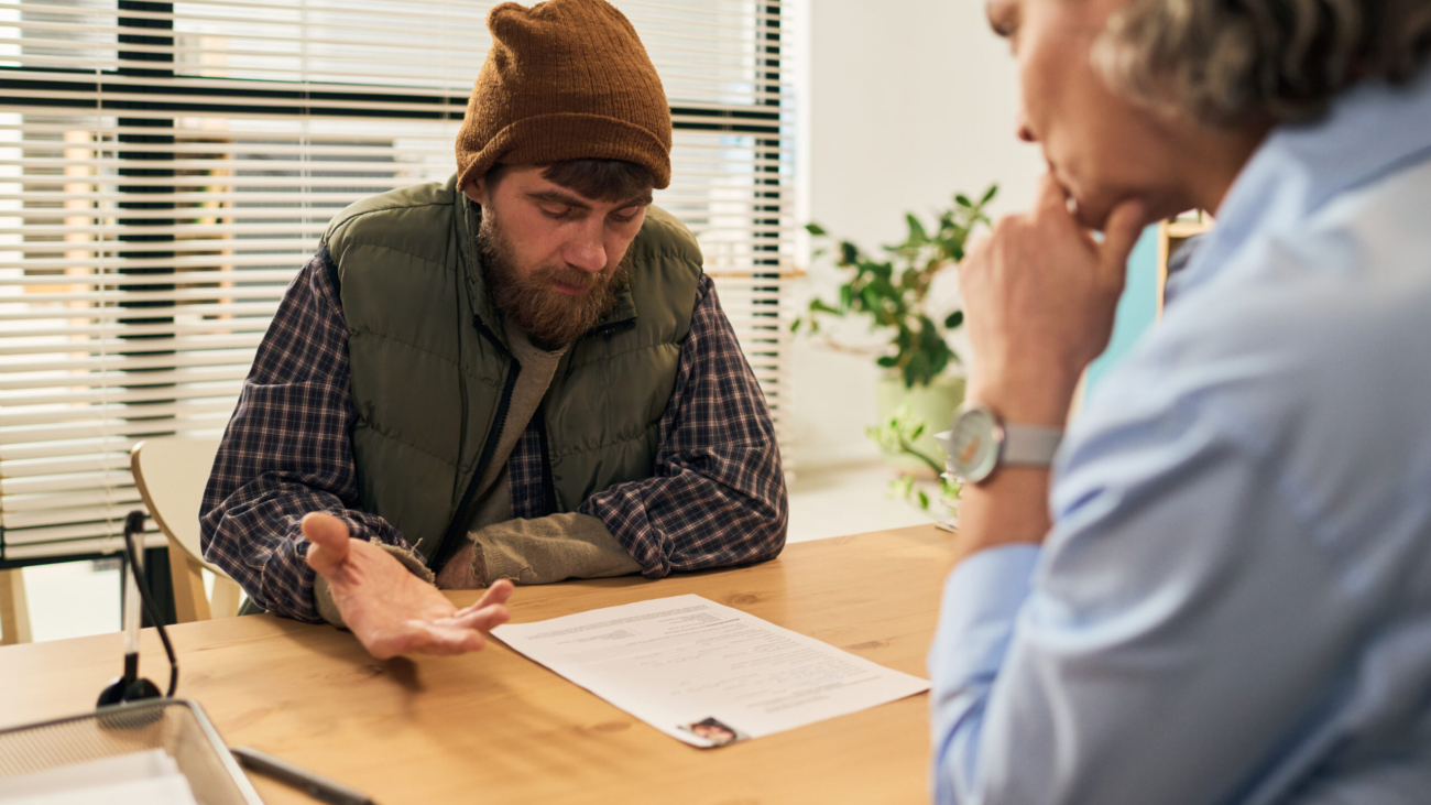 Middle aged Caucasian man wearing beanie and vest discussing paperwork with middle aged Caucasian female social worker in office setting, focusing on social services support