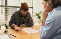 Middle aged Caucasian man wearing beanie and vest discussing paperwork with middle aged Caucasian female social worker in office setting, focusing on social services support